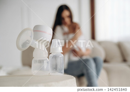 Breast pump and baby bottles placed on a table in a softly lit living room with a mother holding her infant in the background Breast pump and baby bottles placed on a table in a softly lit living room with a mother holding her infant in the background 142451840