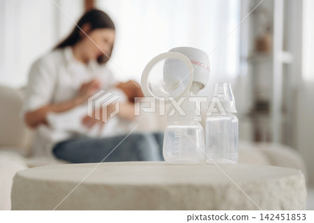 Mother using breast pump with baby in her arms, two baby bottles placed on a round table in a bright, cozy living room setting 142451853