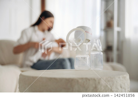 Breast pump and baby bottles on a table in a cozy living room with a mother breastfeeding her infant on a sofa in the background Breast pump and baby bottles on a table in a cozy living room with a mother breastfeeding her infant on a sofa in the background 142451856