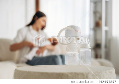 Breast pump and baby bottles placed on a table in a cozy living room with a mother holding her infant in the background, soft natural light illuminating the scene Breast pump and baby bottles placed on a table in a cozy living room with a mother holding her infant in the background, soft natural light illuminating the scene 142451857