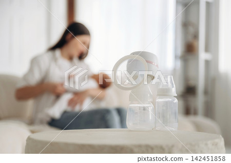 Breast pump and baby bottles placed on a table with a mother breastfeeding her infant in a softly lit living room setting Breast pump and baby bottles placed on a table with a mother breastfeeding her infant in a softly lit living room setting 142451858