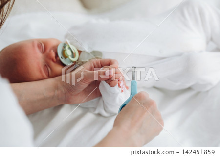 Infant sleeping peacefully in white onesie while caregiver gently trims nails with blue scissors, soft bedding and neutral colors in the background Infant sleeping peacefully in white onesie while caregiver gently trims nails with blue scissors, soft bedding and neutral colors in the background 142451859