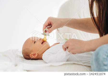 Infant lying on a soft surface while a caregiver gently places a pacifier in the baby's mouth, showcasing a nurturing interaction in a bright indoor setting Infant lying on a soft surface while a caregiver gently places a pacifier in the baby's mouth, showcasing a nurturing interaction in a bright indoor setting 142451874