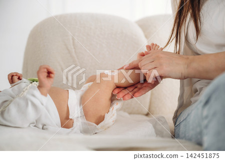Caucasian woman applying cream to the leg of an infant on a soft couch in a bright indoor setting, showcasing a nurturing moment between caregiver and child 142451875