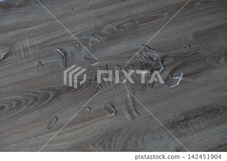 Sharp glass fragments scattered across a wood laminate floor creating a hazardous mess Sharp glass fragments scattered across a wood laminate floor creating a hazardous mess 142451904