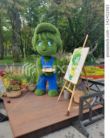 Green topiary boy artist sculpture holding a paint palette near an easel with a canvas picture in a city park flower exhibition Green topiary boy artist sculpture holding a paint palette near an easel with a canvas picture in a city park flower exhibition 142452083