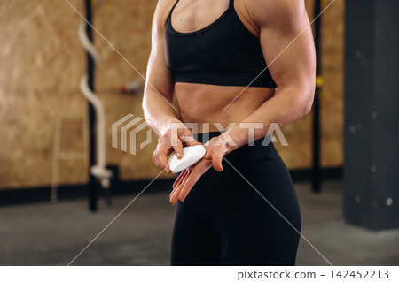 Female athlete applying chalk to hands before workout in a gym setting with wooden walls and exercise equipment visible in the background Female athlete applying chalk to hands before workout in a gym setting with wooden walls and exercise equipment visible in the background 142452213