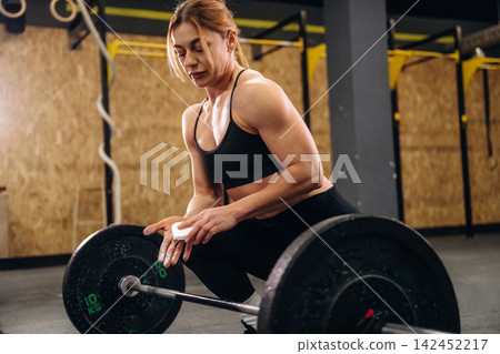 Female athlete in black sportswear prepares to lift a barbell with weights in a gym setting, wooden wall and exercise equipment visible in the background Female athlete in black sportswear prepares to lift a barbell with weights in a gym setting, wooden wall and exercise equipment visible in the background 142452217