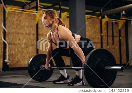 Female athlete performing deadlift exercise with barbell in gym, focused posture, black workout attire, wooden wall and fitness equipment in the background Female athlete performing deadlift exercise with barbell in gym, focused posture, black workout attire, wooden wall and fitness equipment in the background 142452219