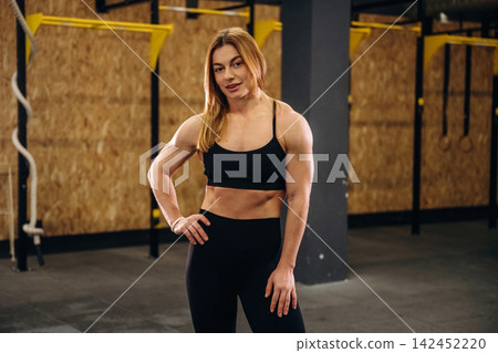 Female athlete in black sportswear poses confidently in a gym setting with wooden walls and fitness equipment visible in the background Female athlete in black sportswear poses confidently in a gym setting with wooden walls and fitness equipment visible in the background 142452220