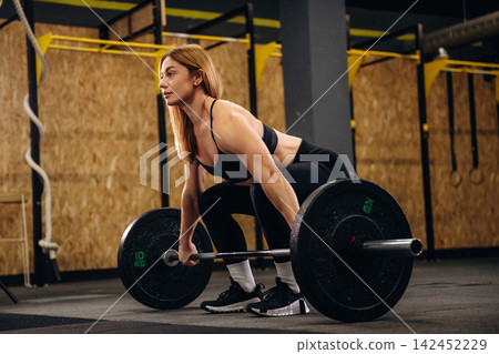 Female weightlifter in black athletic wear prepares to lift a barbell with weights in a gym setting, wooden wall and exercise equipment visible in the background Female weightlifter in black athletic wear prepares to lift a barbell with weights in a gym setting, wooden wall and exercise equipment visible in the background 142452229