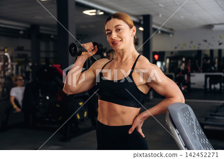 Female fitness enthusiast in black sportswear poses confidently with a dumbbell in a modern gym, exercise equipment and mirrors visible in the background Female fitness enthusiast in black sportswear poses confidently with a dumbbell in a modern gym, exercise equipment and mirrors visible in the background 142452271