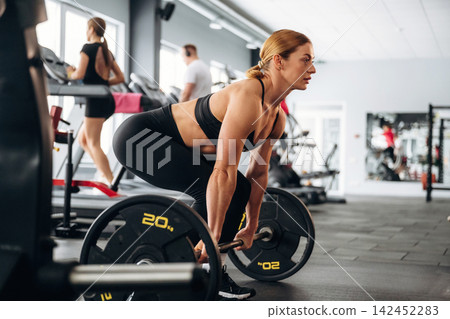 Female athlete performing deadlift exercise with weights in modern gym, fitness equipment and other gym-goers visible in the background Female athlete performing deadlift exercise with weights in modern gym, fitness equipment and other gym-goers visible in the background 142452283