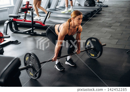 Female athlete performing deadlift exercise with barbell in modern gym, fitness equipment and other gym-goers visible in the background Female athlete performing deadlift exercise with barbell in modern gym, fitness equipment and other gym-goers visible in the background 142452285
