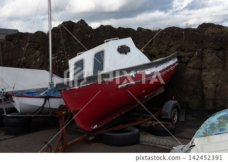 Red fishing boat with a white cabin parked on a trailer by dark rocky cliffs under cloudy sky 142452391