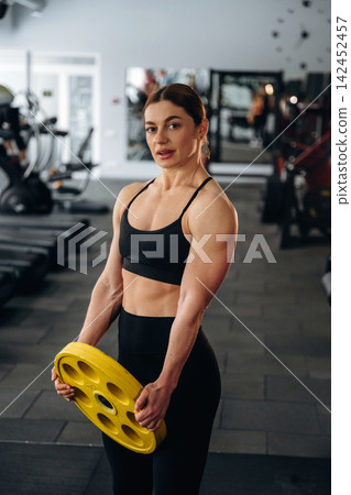Female athlete in black sportswear holds yellow weight plate in gym, fitness equipment and mirrors visible in the background Female athlete in black sportswear holds yellow weight plate in gym, fitness equipment and mirrors visible in the background 142452457