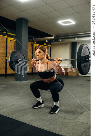 Female athlete performing barbell squats in a modern gym with weight plates on the bar and fitness equipment visible in the background Female athlete performing barbell squats in a modern gym with weight plates on the bar and fitness equipment visible in the background 142452472