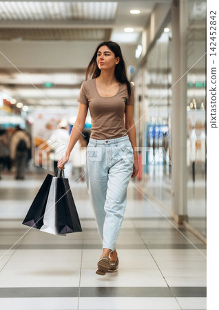 Young woman with long dark hair wearing a brown shirt and light blue pants walks through a shopping mall carrying black and white shopping bags in her hand Young woman with long dark hair wearing a brown shirt and light blue pants walks through a shopping mall carrying black and white shopping bags in her hand 142452842