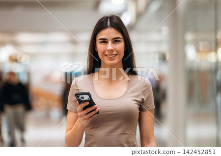 Young woman with long dark hair holding smartphone smiles while standing in a modern shopping mall with blurred shoppers in the background Young woman with long dark hair holding smartphone smiles while standing in a modern shopping mall with blurred shoppers in the background 142452851