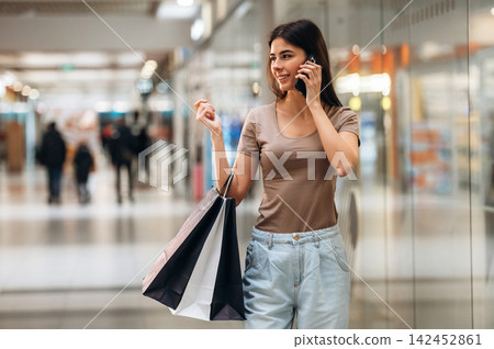 Young woman talking on smartphone while holding shopping bags in a modern shopping mall with blurred shoppers and store displays in the background Young woman talking on smartphone while holding shopping bags in a modern shopping mall with blurred shoppers and store displays in the background 142452861