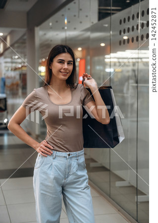Young woman with long dark hair wearing a beige top and light blue jeans holds a shopping bag while standing in a modern shopping mall interior Young woman with long dark hair wearing a beige top and light blue jeans holds a shopping bag while standing in a modern shopping mall interior 142452871