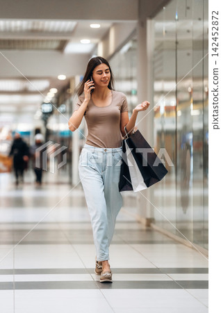 Young woman walking in a shopping mall, talking on her phone while carrying shopping bags, with store reflections visible in the background 142452872