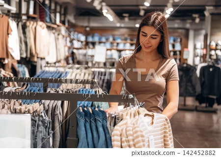 Young woman shopping for clothes in a retail store, examining a striped shirt while surrounded by various garments on display racks in a modern clothing store Young woman shopping for clothes in a retail store, examining a striped shirt while surrounded by various garments on display racks in a modern clothing store 142452882