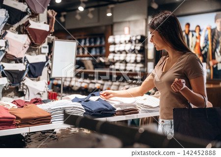 Young woman shopping for clothing in a retail store, examining folded garments on a display table with organized shelves of apparel in the background Young woman shopping for clothing in a retail store, examining folded garments on a display table with organized shelves of apparel in the background 142452888