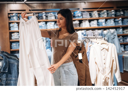 Young woman examining white pants while shopping in a clothing store, denim garments displayed on shelves in the background, casual fashion retail environment Young woman examining white pants while shopping in a clothing store, denim garments displayed on shelves in the background, casual fashion retail environment 142452917