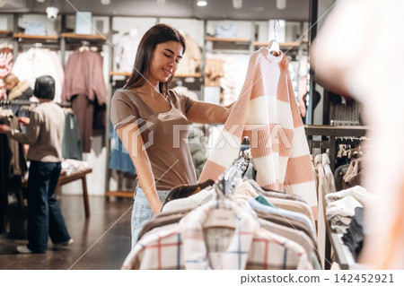 Young woman shopping for clothing in a retail store, holding a striped sweater while browsing through a rack of shirts, with other shoppers visible in the background Young woman shopping for clothing in a retail store, holding a striped sweater while browsing through a rack of shirts, with other shoppers visible in the background 142452921
