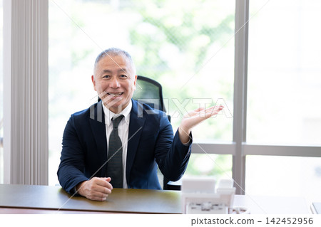 一位資深商人面帶微笑,雙手在辦公桌前張開,一邊指揮一邊發言。 一位資深商人面帶微笑,雙手在辦公桌前張開,一邊指揮一邊發言。 142452956