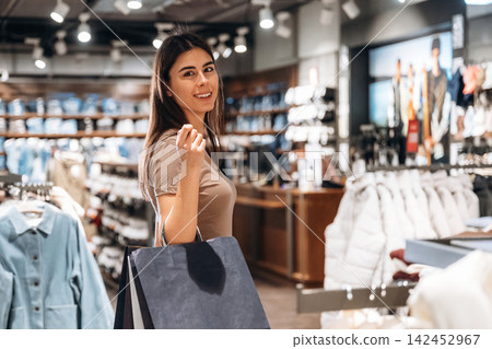Young woman with long dark hair smiles while holding shopping bag inside a modern retail clothing store with various garments displayed around her Young woman with long dark hair smiles while holding shopping bag inside a modern retail clothing store with various garments displayed around her 142452967