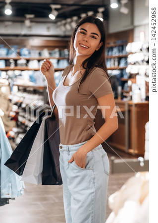 Young woman with long dark hair smiles while holding shopping bags inside a clothing store, shelves of apparel and accessories visible in the background Young woman with long dark hair smiles while holding shopping bags inside a clothing store, shelves of apparel and accessories visible in the background 142452968