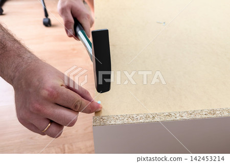 Man holds a nail and swings a hammer at a hardboard back panel Man holds a nail and swings a hammer at a hardboard back panel 142453214