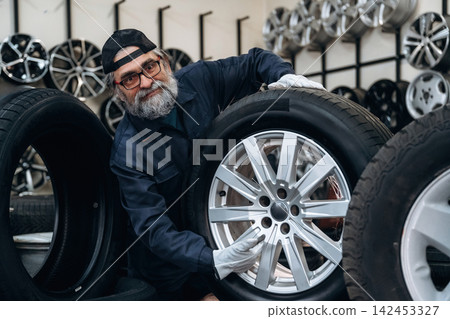 Senior male technician with glasses inspects a silver alloy wheel while surrounded by various tires in a tire shop with a display of rims in the background Senior male technician with glasses inspects a silver alloy wheel while surrounded by various tires in a tire shop with a display of rims in the background 142453327