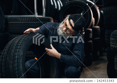 Elderly Caucasian male mechanic inspects a tire in a tire shop surrounded by stacks of tires, wearing a blue jumpsuit and a black cap, focused on the task at hand 142453343