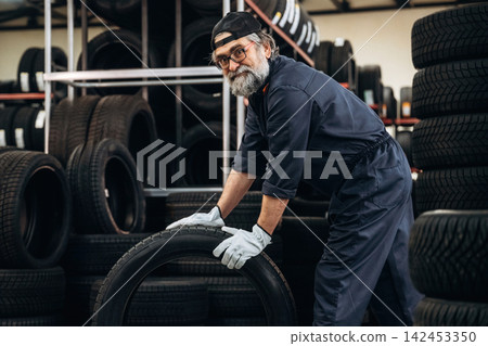 Senior male mechanic in blue coveralls lifts a tire in a tire storage facility surrounded by stacks of tires and a well-organized workshop environment 142453350