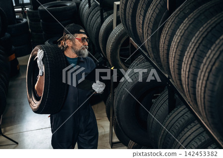 Caucasian male tire technician wearing gloves inspects tire while holding clipboard in tire storage area with stacked tires in the background Caucasian male tire technician wearing gloves inspects tire while holding clipboard in tire storage area with stacked tires in the background 142453382