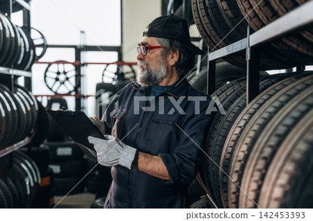 Senior male mechanic wearing glasses and gloves inspects tire inventory while holding a clipboard in a tire shop with shelves of tires in the background Senior male mechanic wearing glasses and gloves inspects tire inventory while holding a clipboard in a tire shop with shelves of tires in the background 142453393