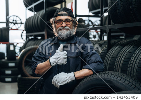 Elderly Caucasian male tire technician wearing gloves poses with thumbs up in a tire shop surrounded by stacks of tires and wheels in the background Elderly Caucasian male tire technician wearing gloves poses with thumbs up in a tire shop surrounded by stacks of tires and wheels in the background 142453409