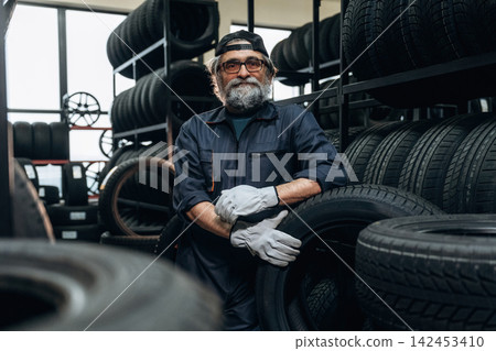 Senior male tire technician wearing gloves poses with a tire in a tire shop, shelves filled with various tires and wheels visible in the background Senior male tire technician wearing gloves poses with a tire in a tire shop, shelves filled with various tires and wheels visible in the background 142453410