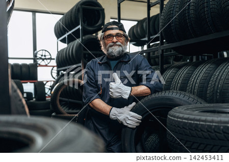 Senior male mechanic in blue coveralls poses with a tire in a tire shop, surrounded by shelves stacked with various tires and wheels in the background Senior male mechanic in blue coveralls poses with a tire in a tire shop, surrounded by shelves stacked with various tires and wheels in the background 142453411