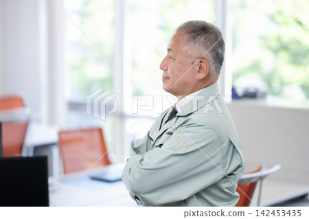 A senior male engineer in work clothes stands with his arms crossed in the office. 142453435