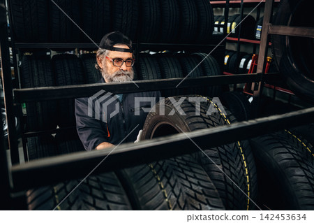 Caucasian male tire technician wearing glasses inspects tires on a rack in an automotive shop, surrounded by stacks of tires and tools in the background Caucasian male tire technician wearing glasses inspects tires on a rack in an automotive shop, surrounded by stacks of tires and tools in the background 142453634