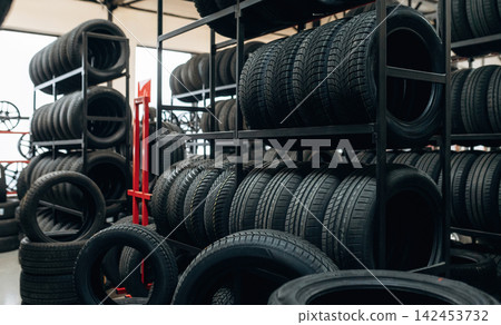 Stack of black rubber tires displayed on metal racks inside a tire shop with additional tires visible in the background and bright overhead lighting illuminating the space Stack of black rubber tires displayed on metal racks inside a tire shop with additional tires visible in the background and bright overhead lighting illuminating the space 142453732