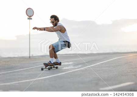 Skateboarder rides along a coastal road during sunset while a person uses a phone nearby 142454308