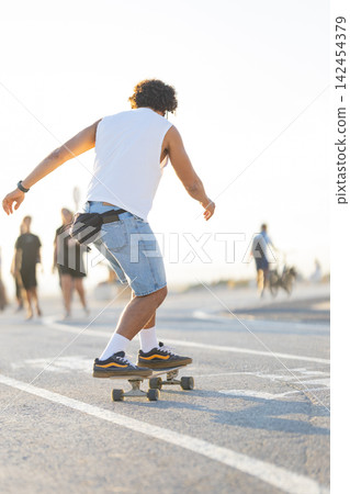 Skateboarder rides along a busy sidewalk during sunset near the beach Skateboarder rides along a busy sidewalk during sunset near the beach 142454379