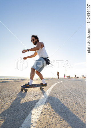 Skateboarder rides along a sunny path near the beach with people in the background during the afternoon Skateboarder rides along a sunny path near the beach with people in the background during the afternoon 142454407
