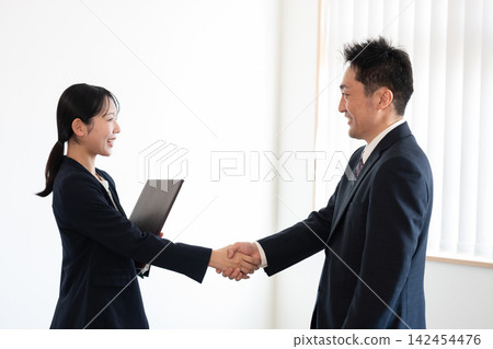 A man and a woman in suits, both business people, exchange smiles and handshakes in an office. A man and a woman in suits, both business people, exchange smiles and handshakes in an office. 142454476