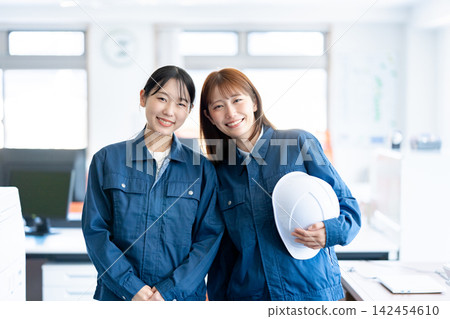 Two smiling female staff members in work clothes, holding helmets, in the office. Two smiling female staff members in work clothes, holding helmets, in the office. 142454610